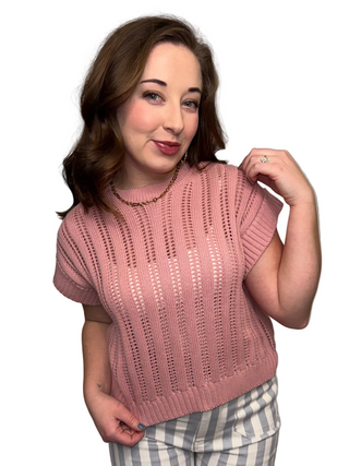 A woman wearing the Rosewater Ribbed Knit in Rose, a dusty pink open-knit cap sleeve sweater with a vertical pointelle pattern. Styled with gray and white striped trousers and a gold necklace.