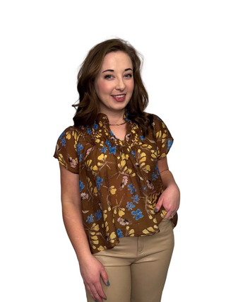 A woman wearing the Golden Petals Top, a honey-brown floral blouse featuring a split neckline with smocked ruffles and short cap sleeves. The botanical print includes sky blue and yellow flowers, styled with tan vegan leather trousers against a white curtain backdrop.