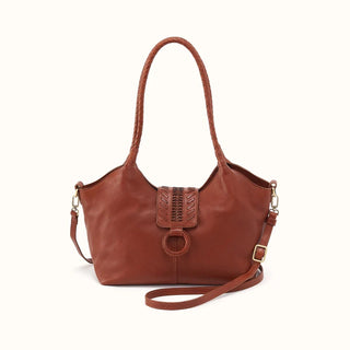 Front view of a slouchy, cognac brown leather shoulder bag presented against a crisp white studio background. The handbag features a distinctive braided leather top handle and a prominent central front tab closure detailed with intricate leather whipstitching and a leather-wrapped O-ring. The smooth leather body is accented by a central vertical seam and includes a secondary adjustable, detachable flat leather crossbody strap secured with gold-tone metal hardware.