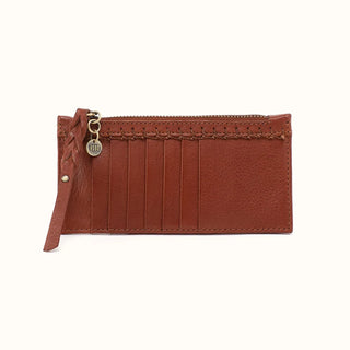 Front view of a HOBO brand brown leather cardholder wallet against a solid white background. The slim, minimalist wallet features a top zipper coin compartment secured by a long, braided leather zipper pull and an antique brass circular charm engraved with the word "HOBO." The exterior face includes six vertical credit card slots positioned below a row of decorative, artisanal whipstitching that spans the width of the accessory, highlighting the rich, natural grain of the brown leather.