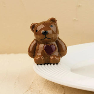 Glossy brown ceramic teddy bear decorative figurine with a dark burgundy heart emblem on its chest, designed as a ceramic mini attachment or rim hugger resting on the edge of a textured white rimmed ceramic serving dish. Set against a warm, neutral beige textured backdrop, perfect for whimsical home decor, table accessories, or collectible kitchen accent displays.