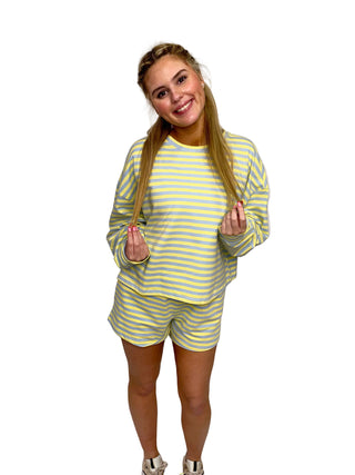 A young woman smiling and holding her hair while wearing the Seaside Sorbet lounge set, featuring a sunshine yellow and sky blue striped French terry sweatshirt and matching relaxed-fit shorts. The outfit is styled with white sneakers against a neutral white curtain backdrop.