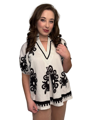A woman wearing the Bohemian Border Blouse, a cream-colored short-sleeve top featuring an intricate bohemian print with blue and earth-toned floral patterns. The blouse has a split V-neckline, short puff sleeves with scalloped blue embroidery, and a relaxed fit, styled against a neutral studio background