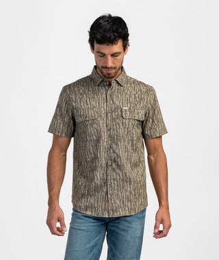 Man posing in the Earthy Oak Tee, a short-sleeve button-down shirt featuring an earthy oak bark vertical camouflage pattern in muted brown and beige tones. The utility-style shirt showcases a structured point collar, a full button placket, double flap chest pockets with button closures, and subtle Western yoke seam details. Styled with medium-wash blue jeans against a plain white studio background.