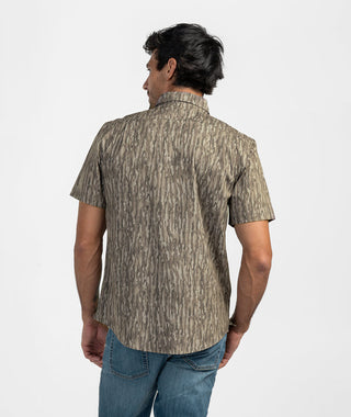 Back view of the Earthy Oak Tee showcasing the full vertical tree bark camouflage design across the back yoke, torso, and short sleeves. Demonstrates the shirt's classic, structured fit and curved hemline worn untucked over blue jeans against a white studio background.