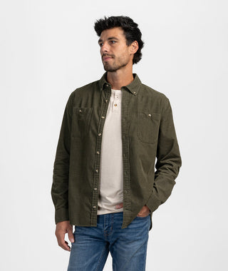 A man posing in an olive green corduroy button-down overshirt worn unbuttoned over an off-white henley shirt and paired with classic blue denim jeans. The shirt features a structured button-down point collar, tortoise-tone buttons, long sleeves with barrel cuffs, and dual front chest patch pockets with button closures, set against a seamless white studio background.