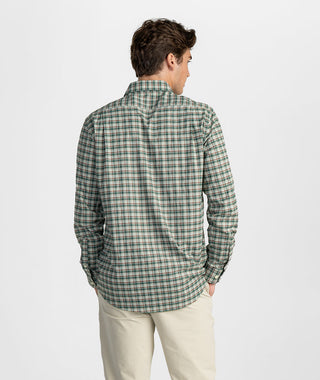 Rear angle of a man wearing a classic-fit long-sleeve button-down shirt featuring an earthy green, brown, and cream plaid tartan pattern. The shirt features a clean back shoulder yoke, full-length set-in sleeves with tailored button cuffs, and a curved shirttail hem. Styled casually untucked over off-white chino trousers against a minimalist studio background.