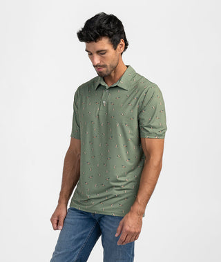 Man posing in the Mallard In Motion Polo, a short-sleeve performance polo shirt in a sage green shade featuring an all-over micro-print of flying mallard ducks. The top showcases a classic structured collar and a three-button placket with light-toned buttons. Styled casually with medium-wash blue jeans against a clean white studio background.