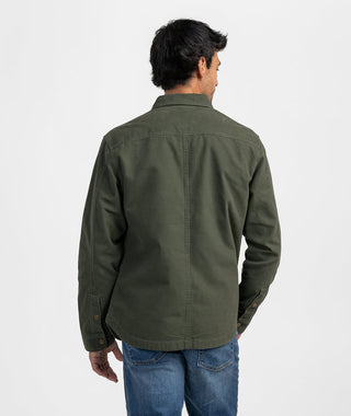 Rear view of a man wearing the olive green utility chore jacket paired with medium-wash blue denim jeans against a seamless white studio backdrop. Highlights the clean yoke seam across the shoulders, reinforced center back vertical stitching, adjustable snap-cuff sleeves, and a curved hem tailored for relaxed, casual layering.