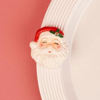 A glossy ceramic Santa Claus mini attachment attached to the fluted rim of a white serving platter or dish. Santa features a cheerful, smiling face with rosy cheeks, closed happy eyes, a classic white beard, and a red Santa hat embellished with a green holly sprig and tiny gold bells. Set against a textured, warm pink backdrop that highlights the festive holiday decor detail.