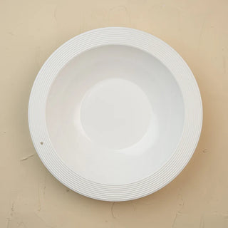 Direct top-down overhead view of a clean, bright white ceramic shallow pasta bowl featuring a wide, ridged accent rim with fine concentric circular grooves. The smooth, glossy interior slopes down gently into a circular base, complete with a small attachment hole on the wide lip designed for interchangeable charms or mini rim toppers. Set against a warm, neutral beige plaster-textured background, ideal for modern table settings, minimalist dinnerware collections, and entertaining displays.
