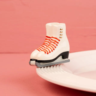 A ceramic decorative mini topper featuring a detailed pair of vintage white figure ice skates with vibrant red painted laces, metallic gold lace hooks, and grey-and-black silver blades. The glossy ceramic piece is attached to the rim of a ribbed white ceramic serving platter against a soft pink backdrop, ideal for seasonal winter table settings and holiday entertaining decor.
